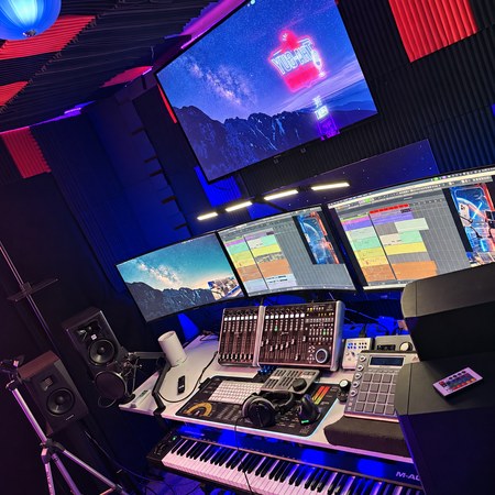 Three desktop monitors below a wall display, music controllers and piano in THA-GUY’s blue and purple studio.