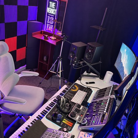White studio chair beside a microphone, speakers, music controllers, piano and the THE VERDICT IS IN neon sign.